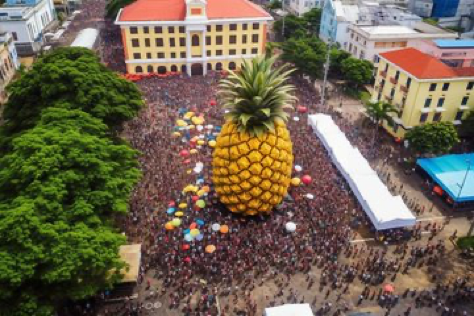 Prompt: ‘’A droneshot of a crowd around the biggest pineapple statue in the world. City square. --ar 3:2’’